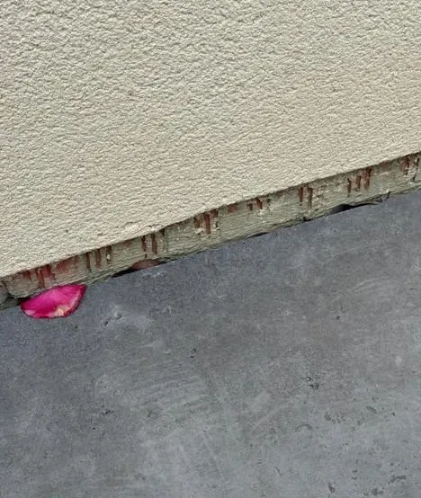 Bas de façade enduite où l'enduit s'arrête et laisse apparaître des briques creuses rouges, sans protection.