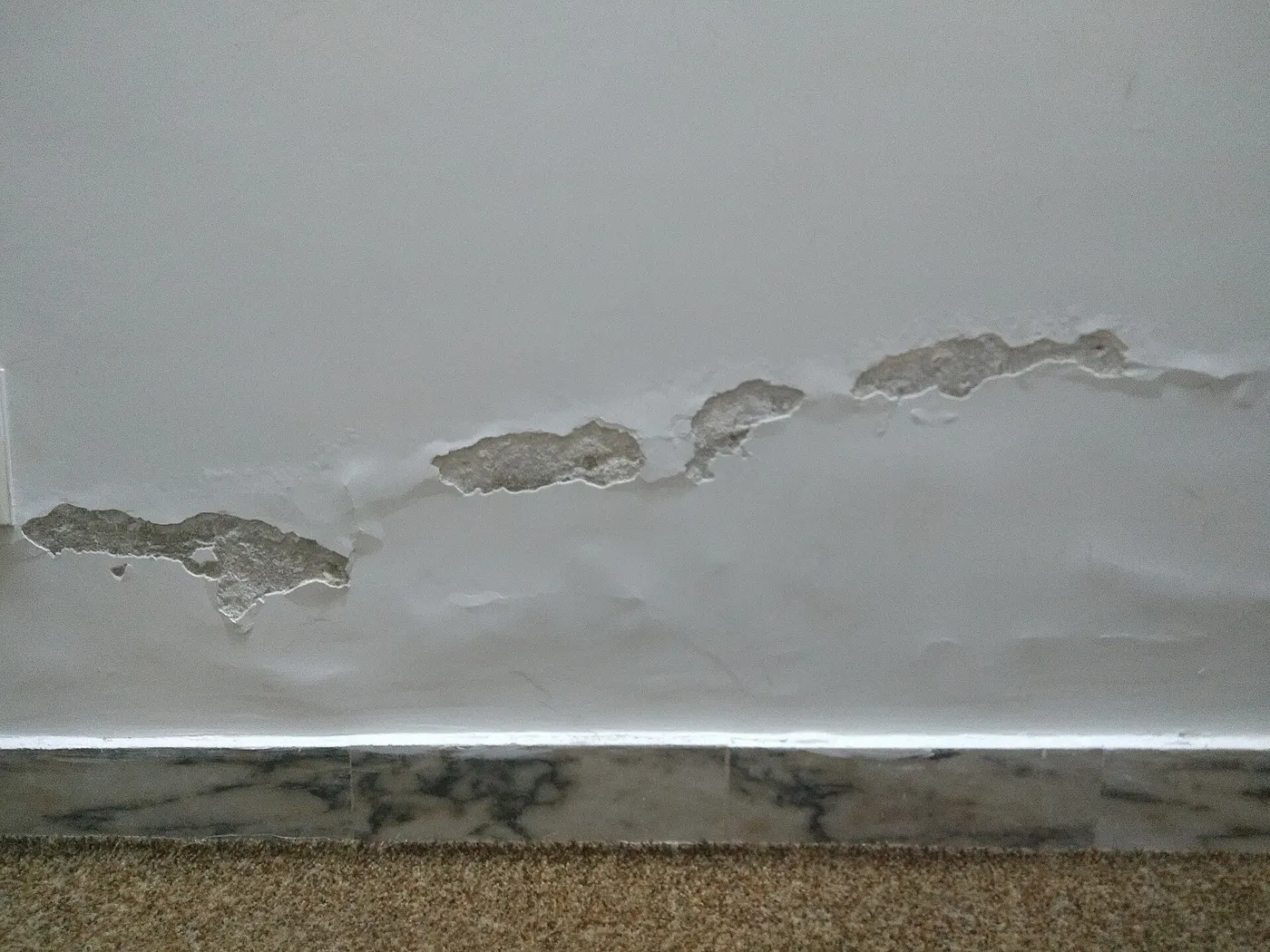 Mur peint dont l'enduit cloque et s'écaille juste au-dessus de la plinthe.