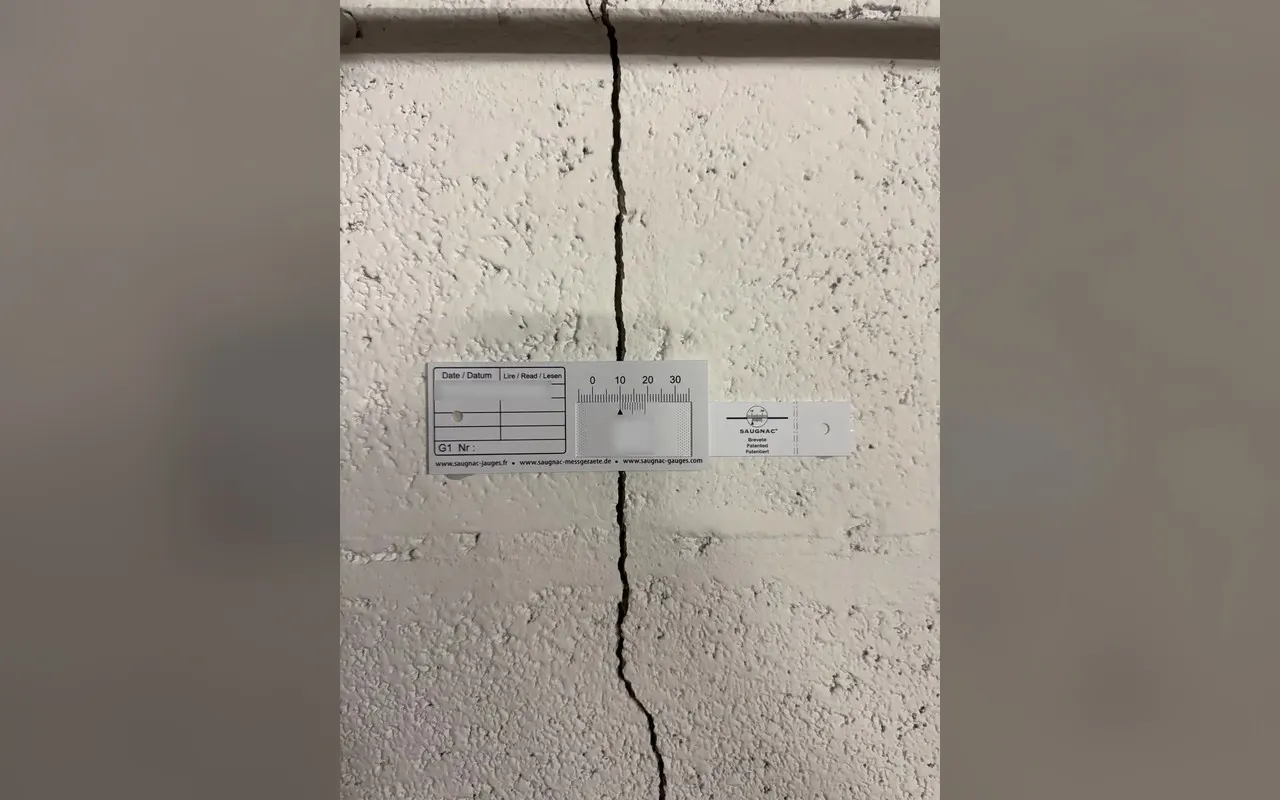 Une jauge posée à cheval sur une fissure verticale dans un enduit rugueux.