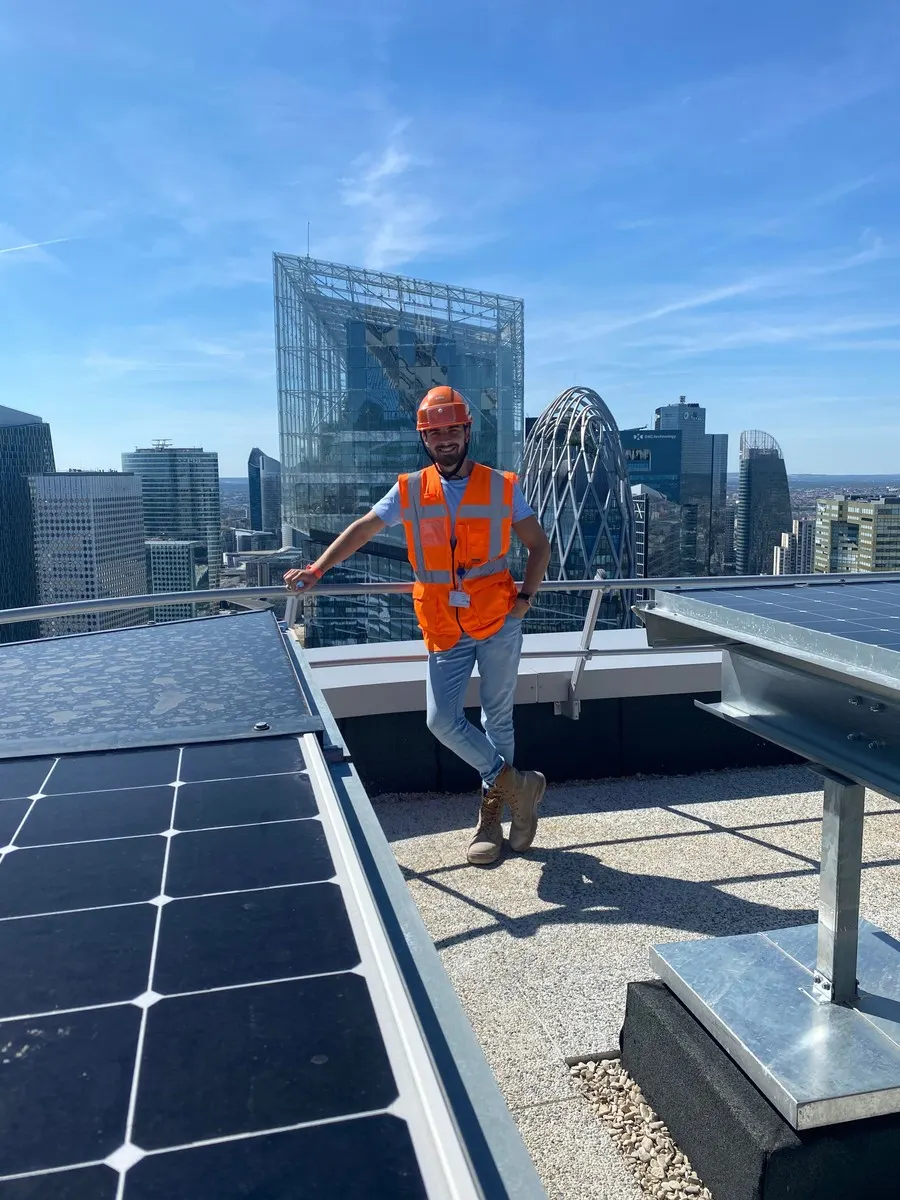 Benoit Gervais, casque et gilet orange de chantier, appuyé à un garde-corps sur le toit de la tour Alto, avec des panneaux solaires au premier plan et les tours de La Défense derrière lui.