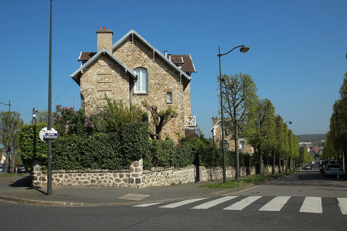 Maison ancienne en pierre meulière, avec ses encadrements et son toit en tuiles, au bord d'une rue bordée d'arbres.