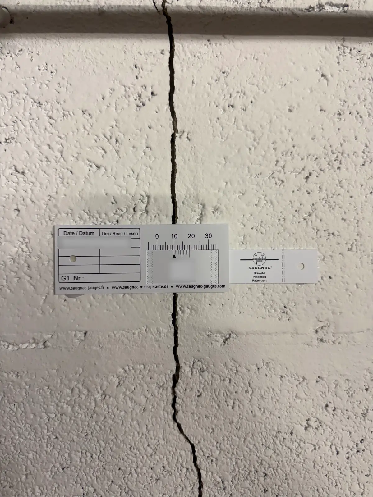 Une jauge graduée posée à cheval sur une fissure verticale dans un mur enduit.