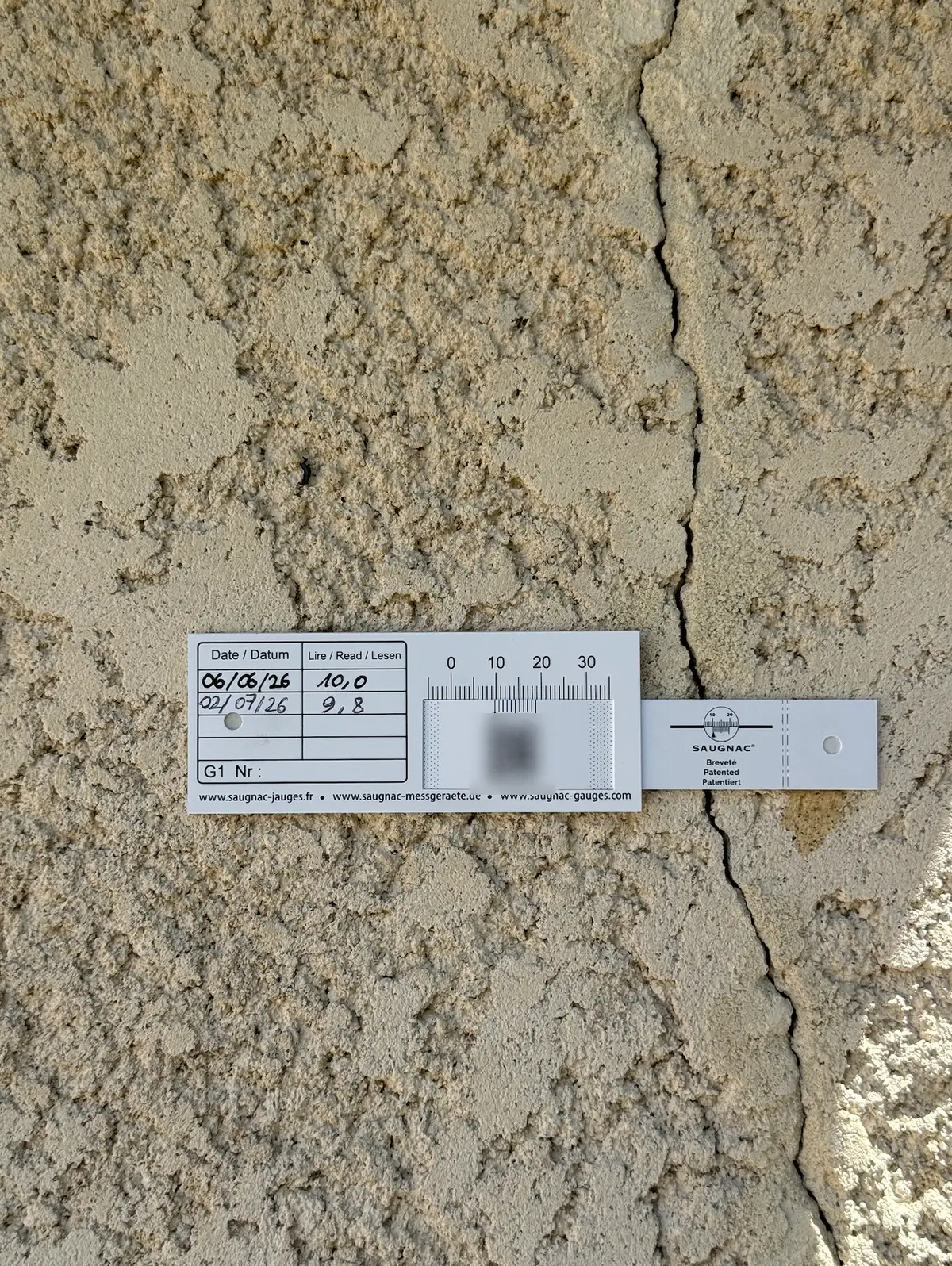 Une jauge fixée sur une fissure dans un enduit, avec deux relevés datés écrits sur l'étiquette.
